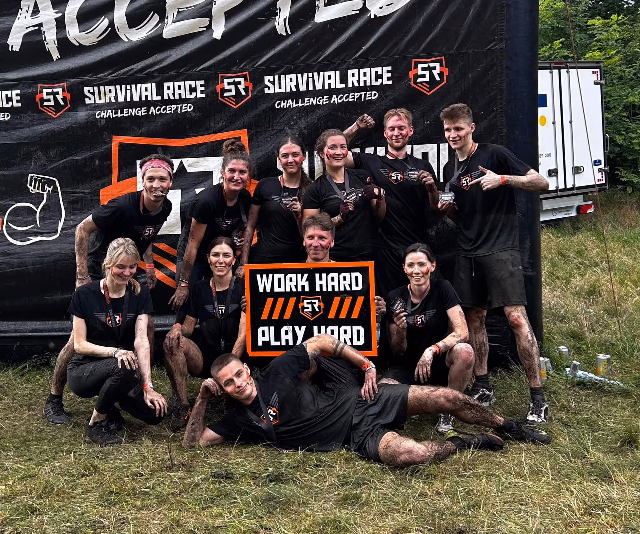 Egala by Boekestijn team after completing a muddy obstacle run, holding a ‘Work Hard Play Hard’ sign — celebrating teamwork, resilience, and strong company spirit.