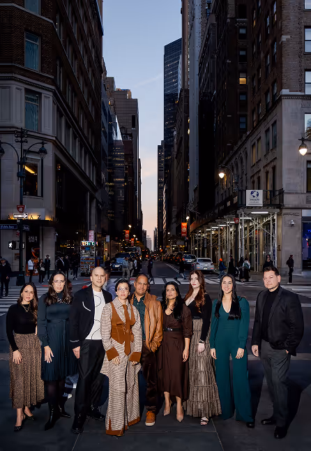Group of eight people dressed in formal and semi-formal attire standing on a city street between tall buildings at dusk.