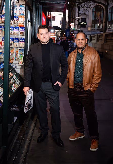 Two men standing on a city sidewalk at night, one in a black suit holding a newspaper and the other in a brown leather jacket.