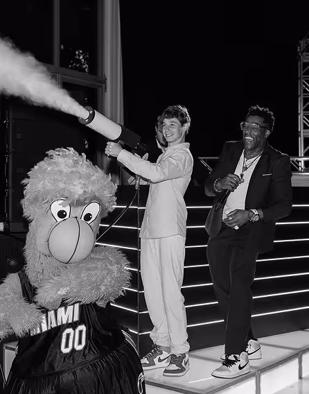 Two men smiling and posing on a lit stage with a Miami Heat mascot spraying smoke from a canister.