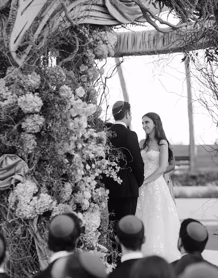 Bride and groom standing under a floral arch during a wedding ceremony with guests wearing yarmulkes in the foreground.