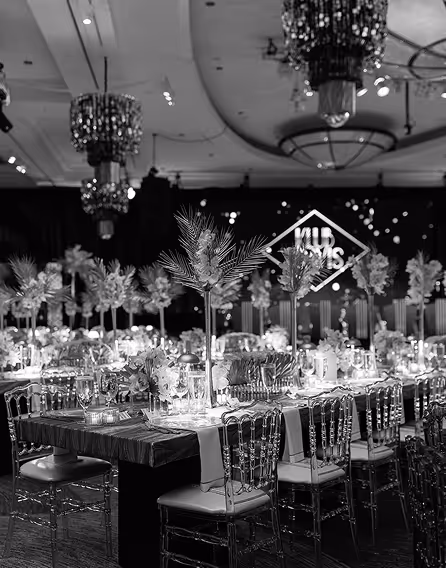 Elegantly decorated banquet hall with tall floral centerpieces on tables and transparent chairs under large chandeliers.