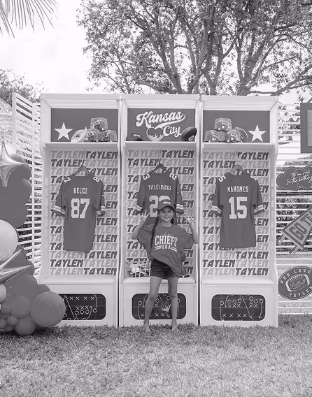 Child wearing a Chiefs t-shirt and cap posing with fists raised in front of display cases featuring Kansas City Chiefs jerseys of Kelce and Mahomes.