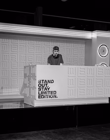Black and white photo of a DJ behind a booth with the text 'STAND OUT. STAY LIMITED EDITION.' on the front panel.