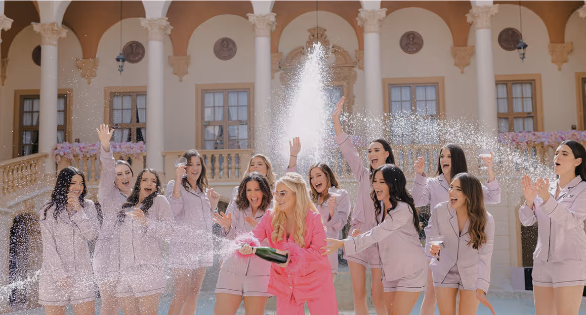 Group of women in matching light purple pajamas celebrating outdoors as one woman in bright pink pajamas sprays champagne.