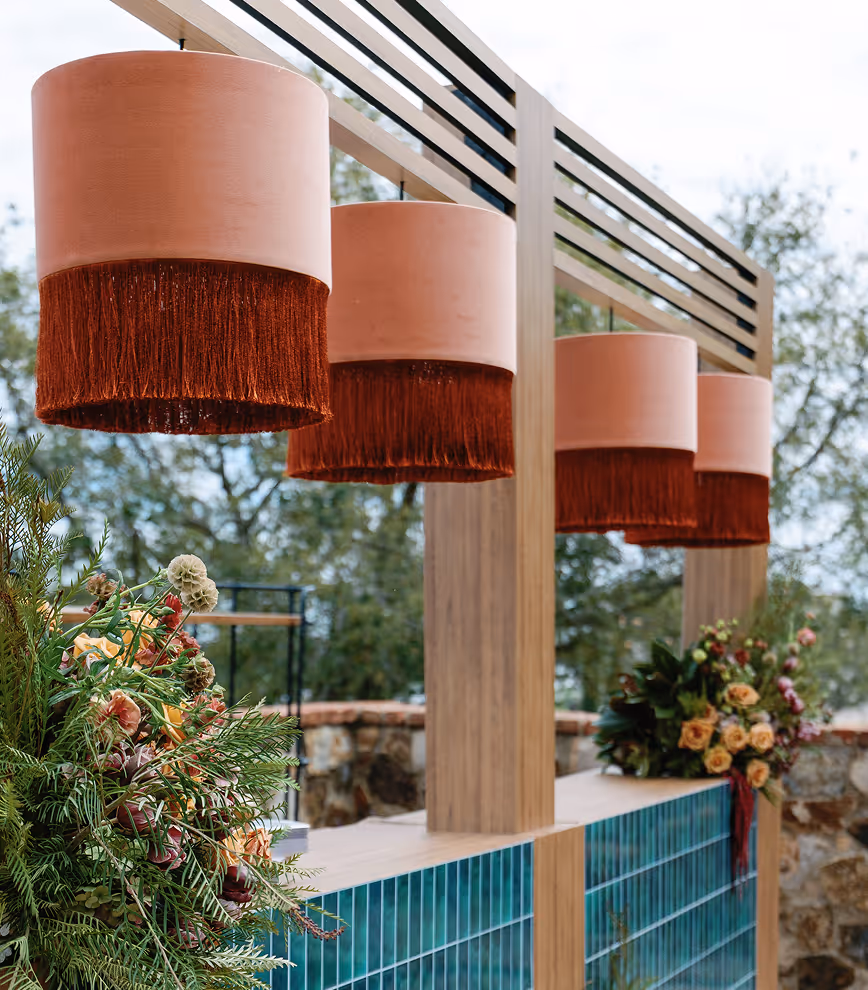 Four cylindrical pendant lamps with beige shades and rust-colored fringe hanging above a bar with teal tiles and floral arrangements.