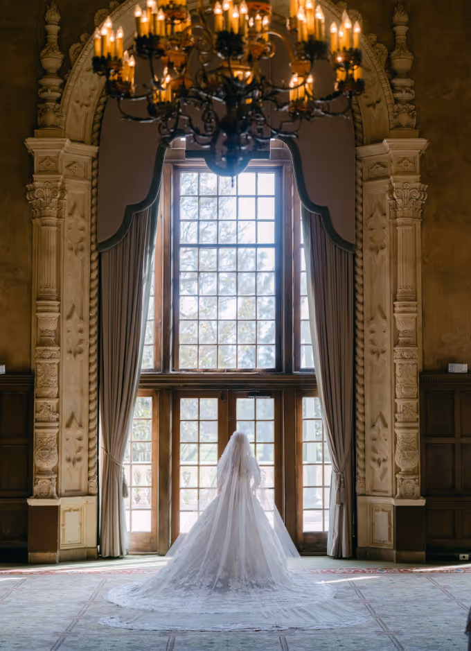 Bride in a long lace wedding gown and veil stands facing large ornate window with curtains in a grand room.