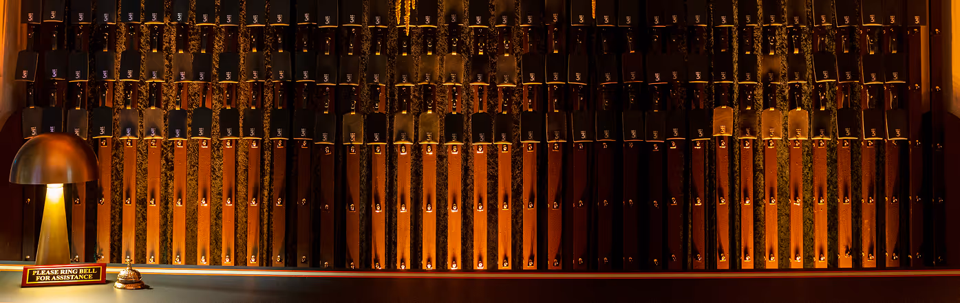 Wall-mounted wooden panel with numerous labeled levers in rows, a lamp, and a bell with a sign reading ‘Please ring bell for assistance.’