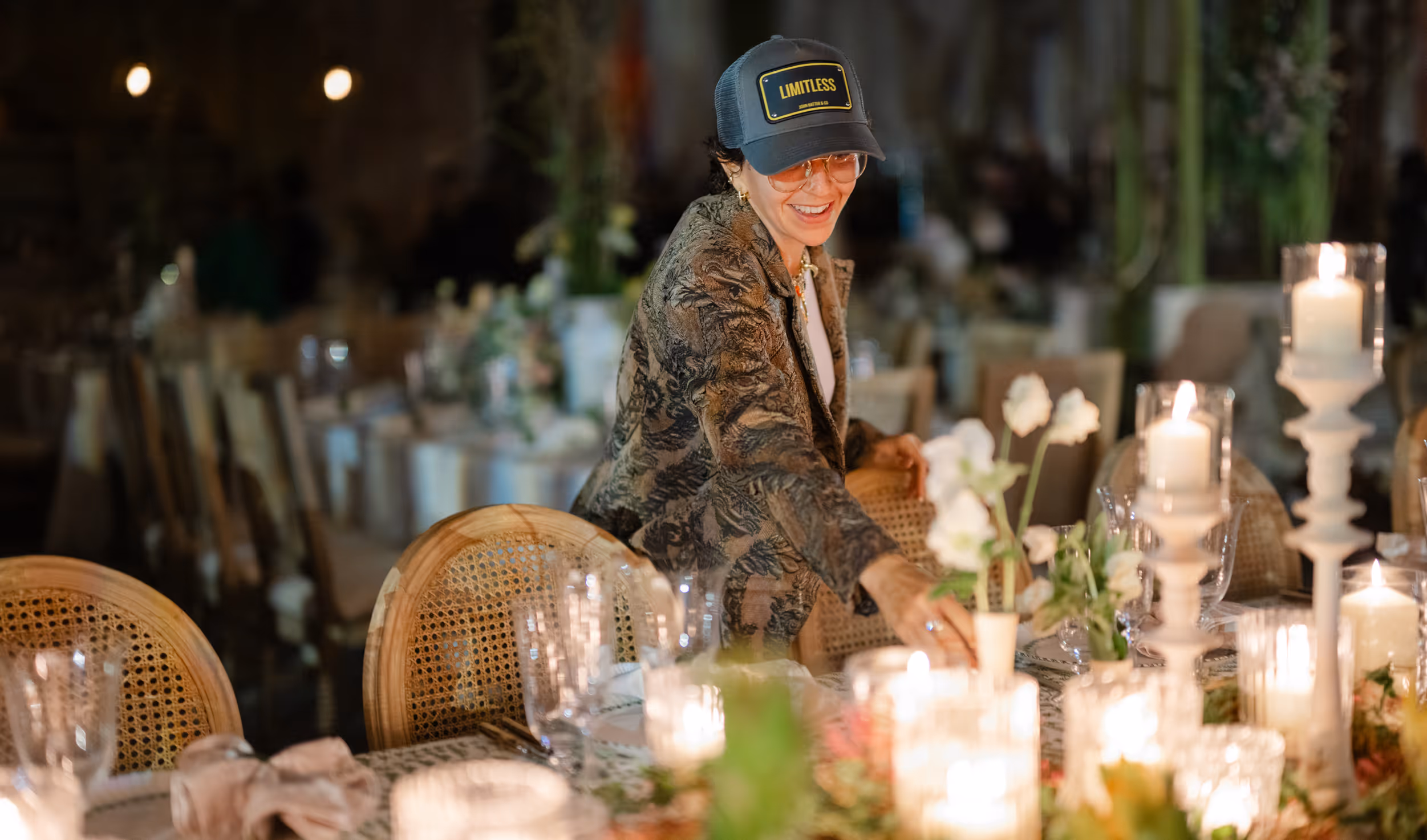 Smiling woman wearing a 'LIMITLESS' cap arranging flowers on a candlelit dining table.