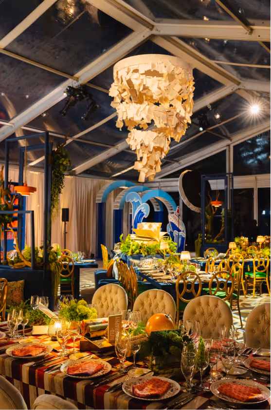 Elegant indoor dining setup with tufted chairs, striped tablecloth, elaborate centerpiece, and a unique hanging chandelier made of paper notes.