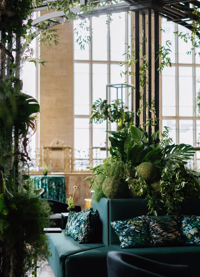 Indoor seating area with green velvet couches, decorative pillows, and lush green plants near large windows.