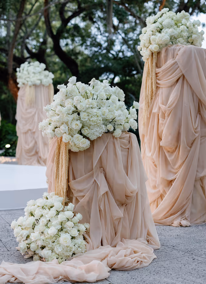 Three beige fabric-draped pedestals adorned with large white floral arrangements outdoors with trees in the background.