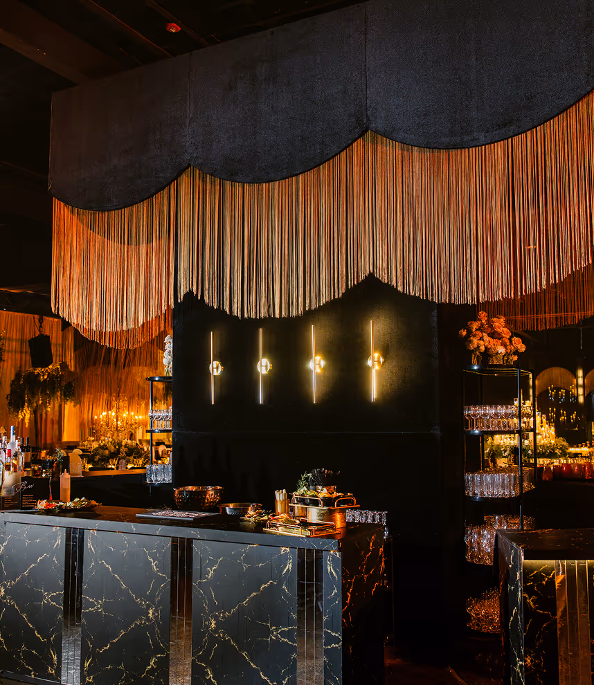 Elegant bar area with black marble counter, vertical gold lighting on black wall, and long gold fringe curtains overhead.