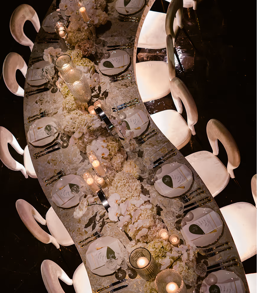 Elegant curved dining table set with white plates, menus, crystal glasses, gold utensils, white floral centerpieces, and lit candles.