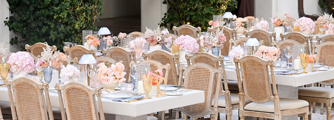 Outdoor dining area with long tables set with decorative glassware, pink and peach floral centerpieces, and vintage wooden chairs.