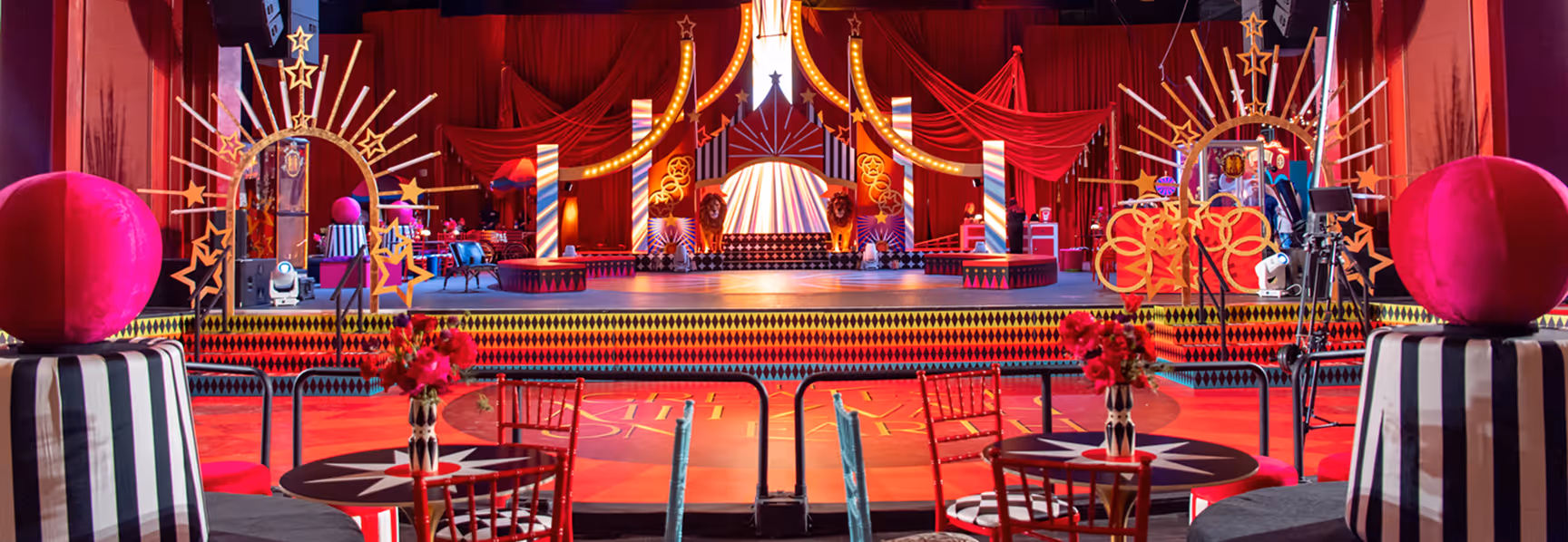 Colorful circus-themed stage with red curtains, decorative stars, lion statues, and tables with striped chairs and floral centerpieces.