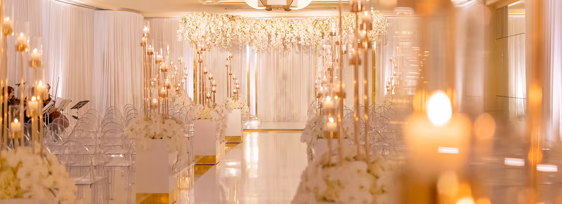 Elegant wedding aisle with white floral arrangements and numerous lit candles in a softly lit venue.