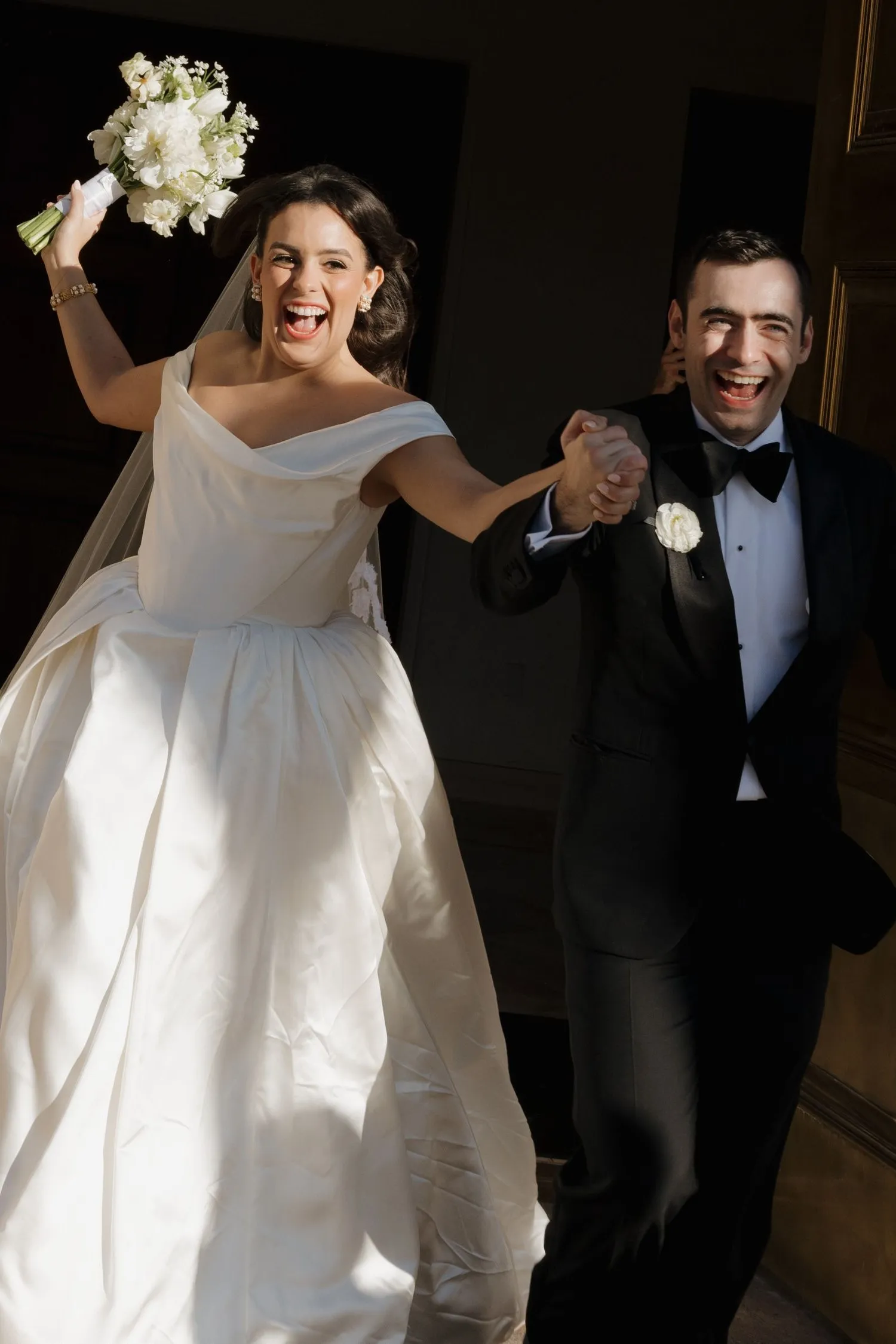 Bride in a white gown holding a bouquet and groom in a black tuxedo holding hands, both smiling joyfully.
