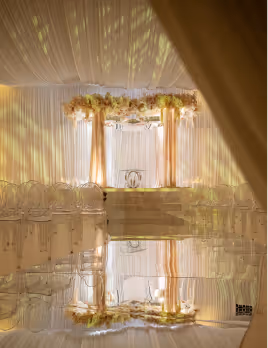 Elegant wedding ceremony setup with floral arch, gold pillars, sheer curtains, and reflective floor.