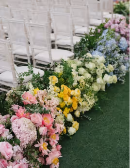 Rows of white chairs lined up beside a green aisle decorated with colorful floral arrangements including pink, yellow, white, and blue flowers.