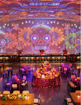 Vibrant event space with colorful Day of the Dead skull projections on the walls and ceiling, decorated tables with floral arrangements and candles.