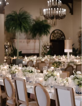 Elegant banquet hall with long dining tables adorned with white floral arrangements, candles, crystal glassware, and upholstered chairs under chandeliers.