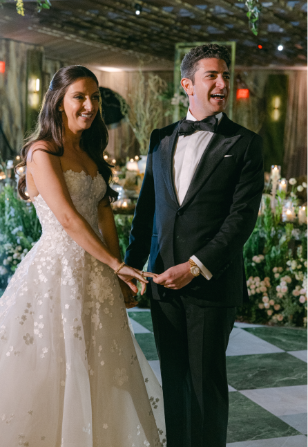Bride in a white floral wedding dress and groom in a black tuxedo holding hands and smiling indoors at a decorated venue.