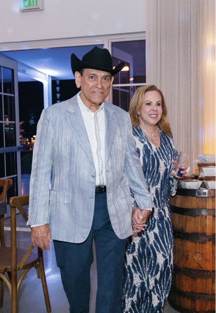 Man in a black cowboy hat and light striped blazer holding hands with a woman in a patterned dress holding a drink, indoors near a wooden barrel and chairs.