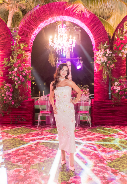 Woman in a strapless white gown standing on a vibrant floral carpet under pink floral arches with a chandelier overhead.