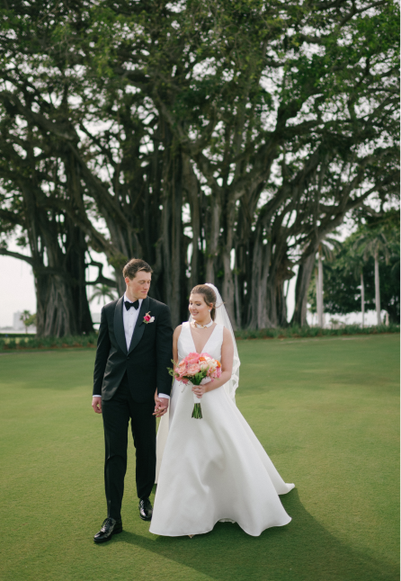 Bride in a white wedding dress holding a pink bouquet and groom in a black tuxedo holding hands walking on green grass with large trees in the background.