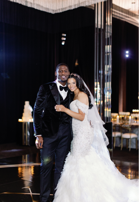 Bride in white wedding gown and groom in black tuxedo smiling and embracing on a dance floor with a wedding cake and decorated reception tables in the background.