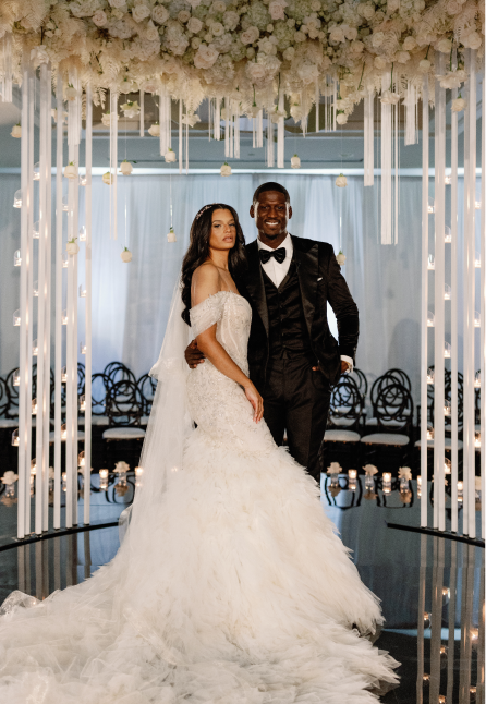 Bride in a white off-shoulder wedding gown and groom in a black tuxedo standing together under a floral ceiling with hanging candles and roses.