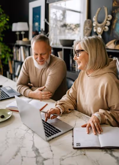 Smiling middle-aged man and woman sitting at a table with a laptop and notebook in a cozy room.