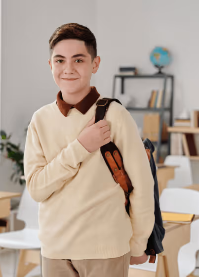 Smiling male student carrying a backpack over one shoulder in a classroom setting with desks and a globe in the background.