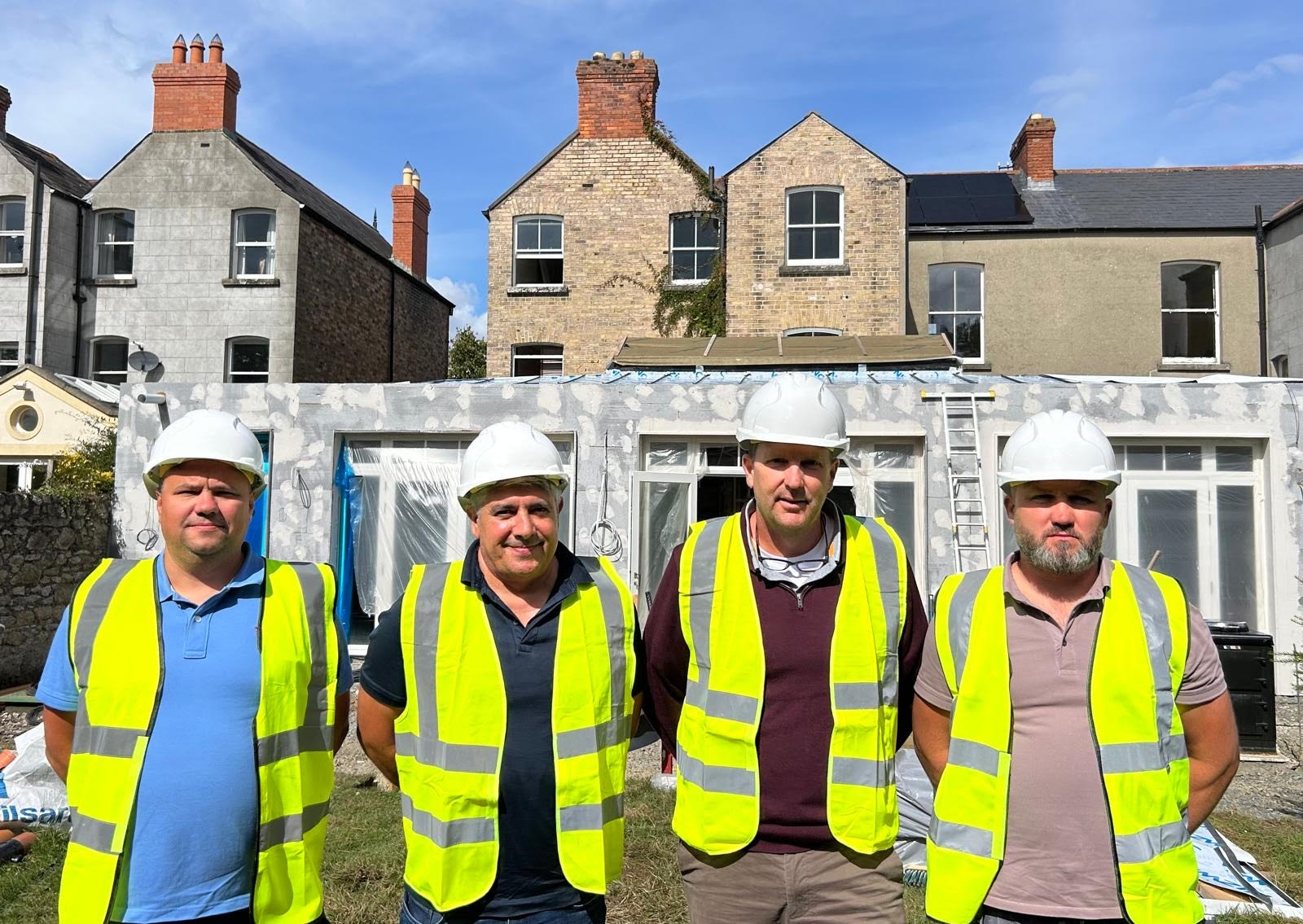 Four construction workers wearing white hard hats and yellow reflective vests standing in front of a partially renovated building with exposed brick houses in the background.