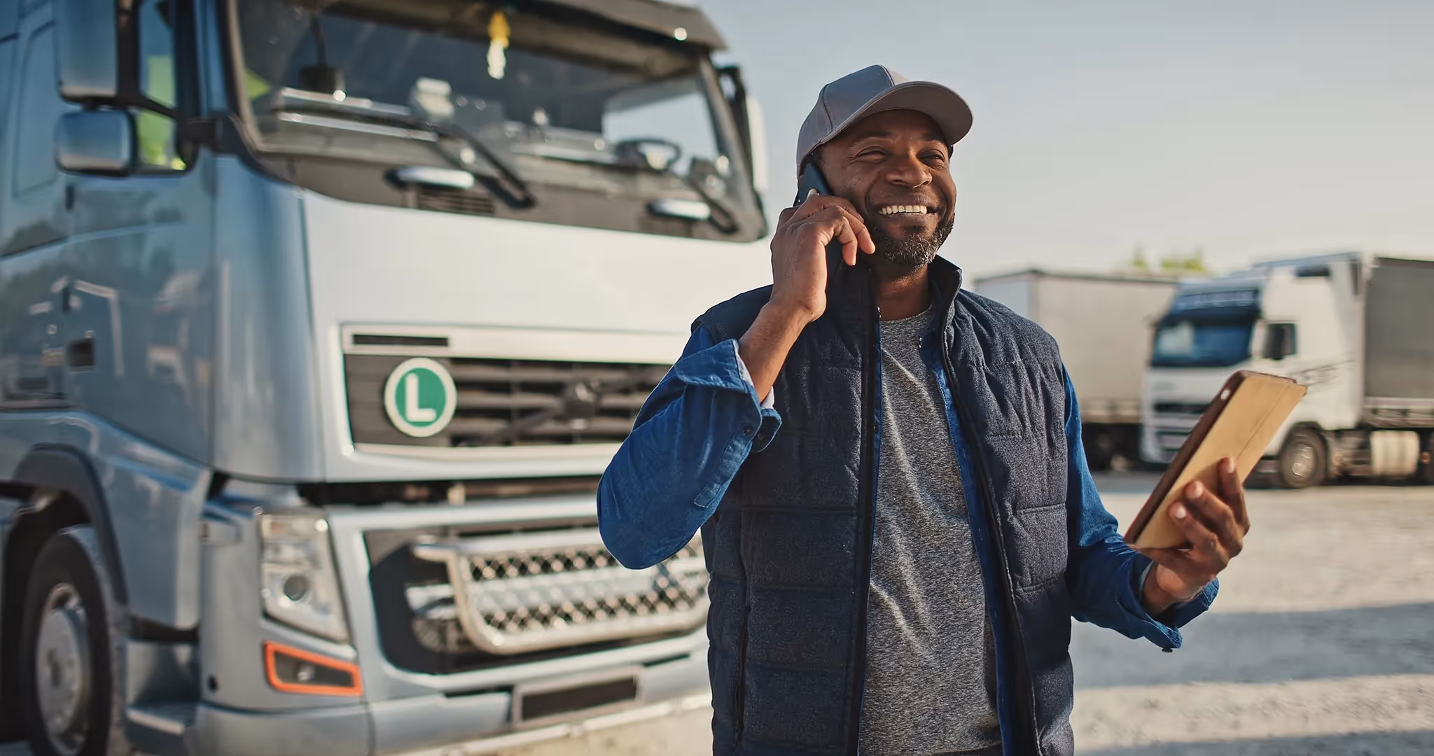 Smiling truck driver standing in front of his commercial vehicle talking on his cellphone and looking at his tablet