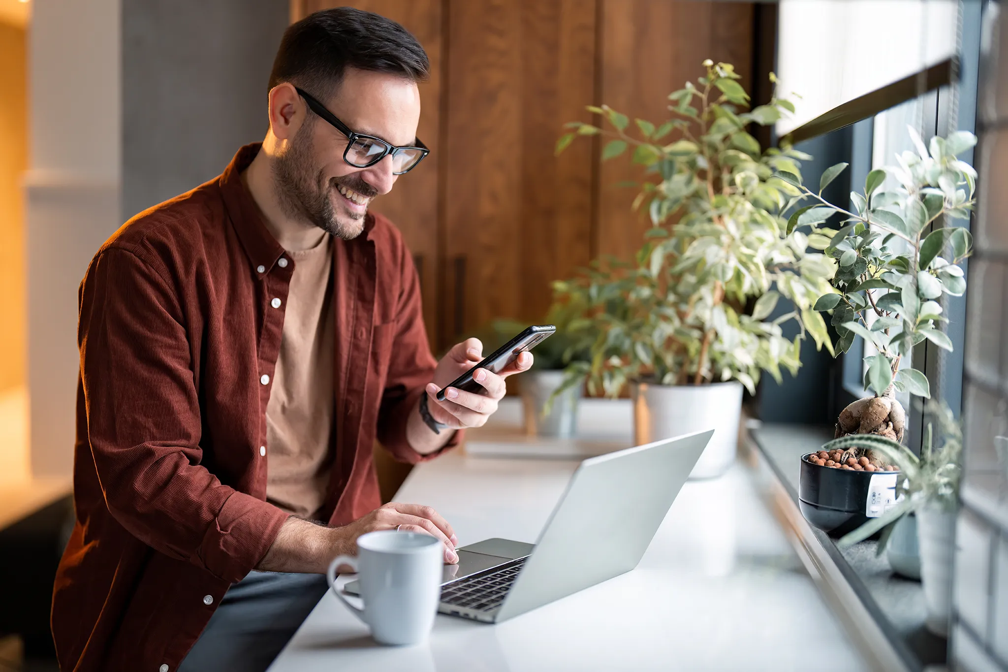 Smiling middle-aged man working at a counter looking at his cellphone and scrolling on his laptop with a coffee