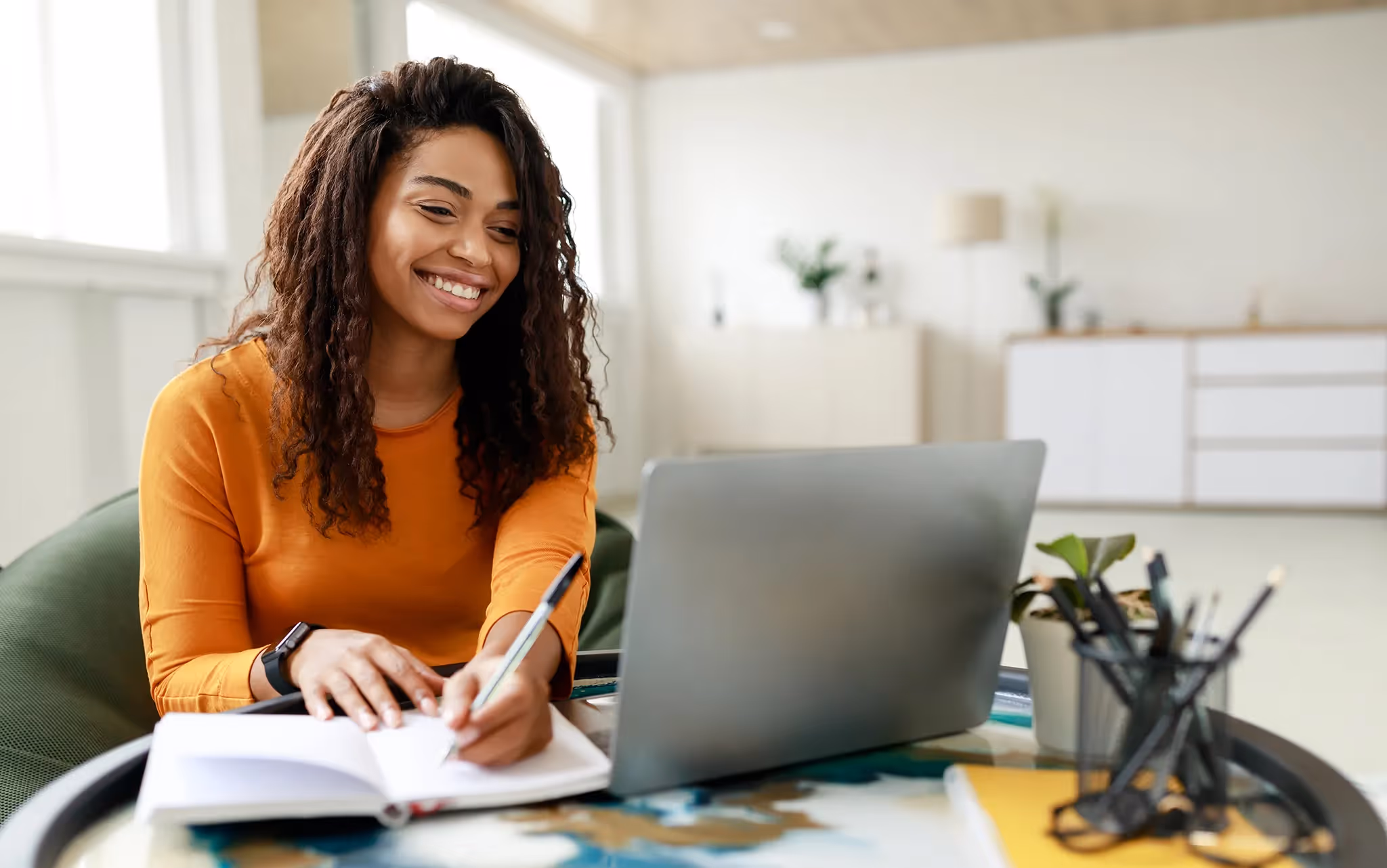 Young woman smiling while looking at her laptop while writing something down in her notebook