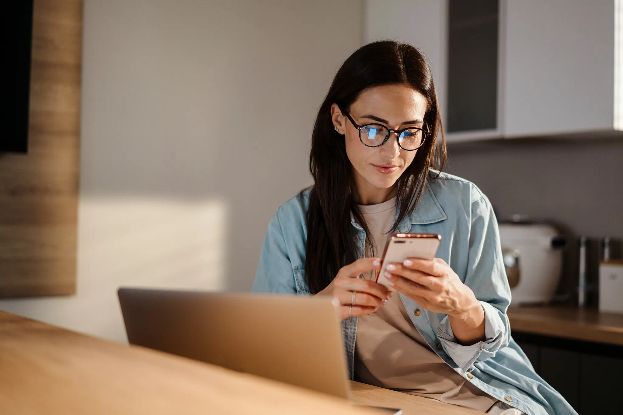 Young woman looking pensively at cellphone while working on laptop