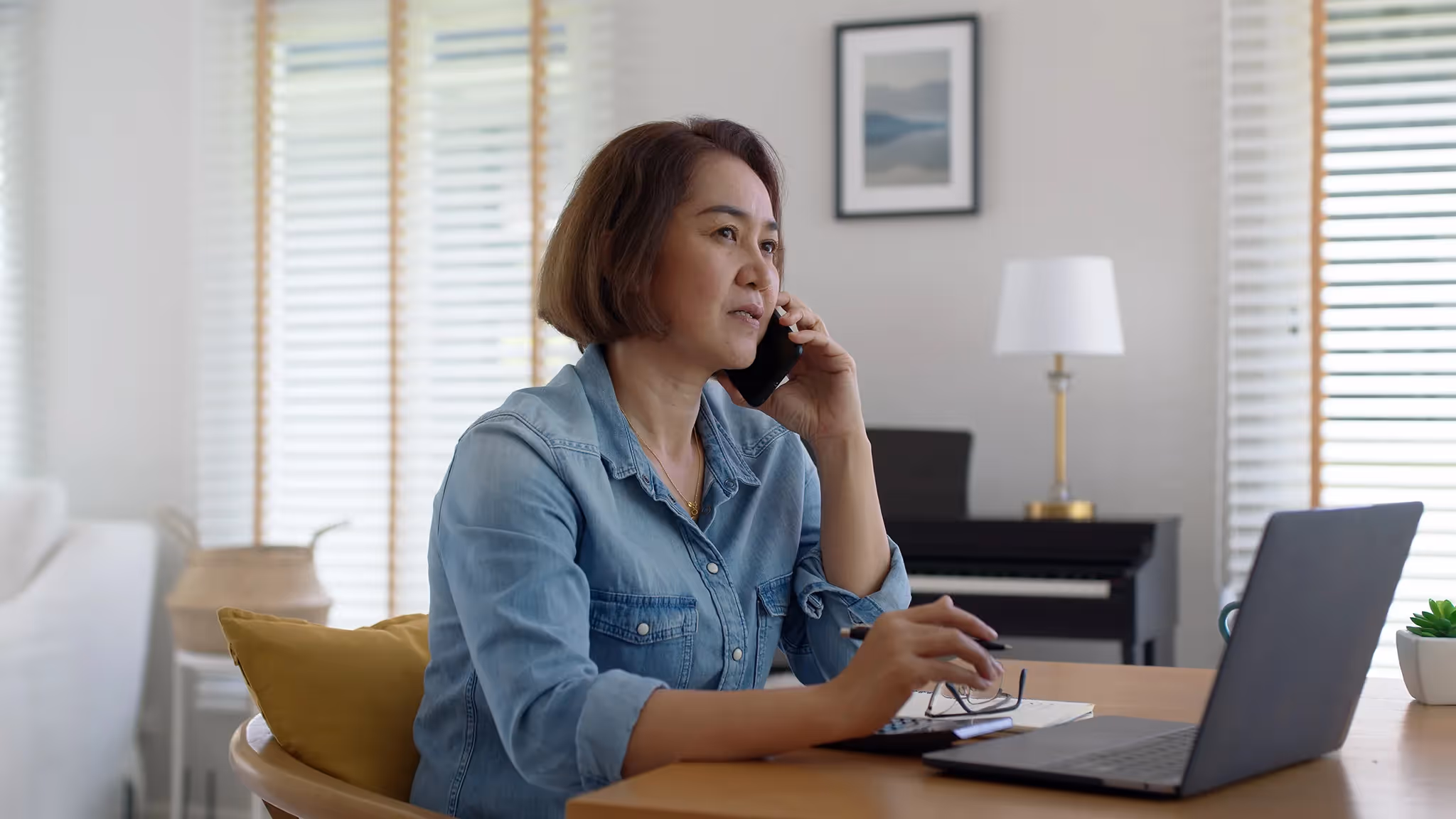 Middle-aged Asian woman researching travel insurance options on her laptop and speaking on a cellphone in her living room