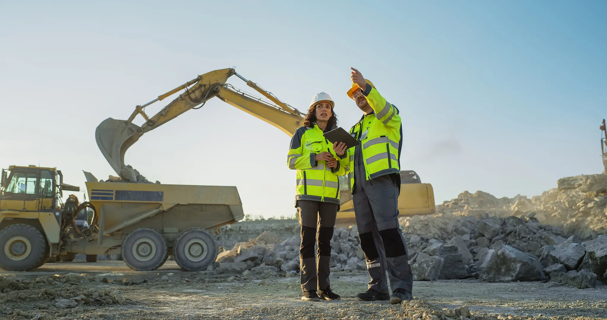 Two construction workers on a jobsite with an excavator in the background