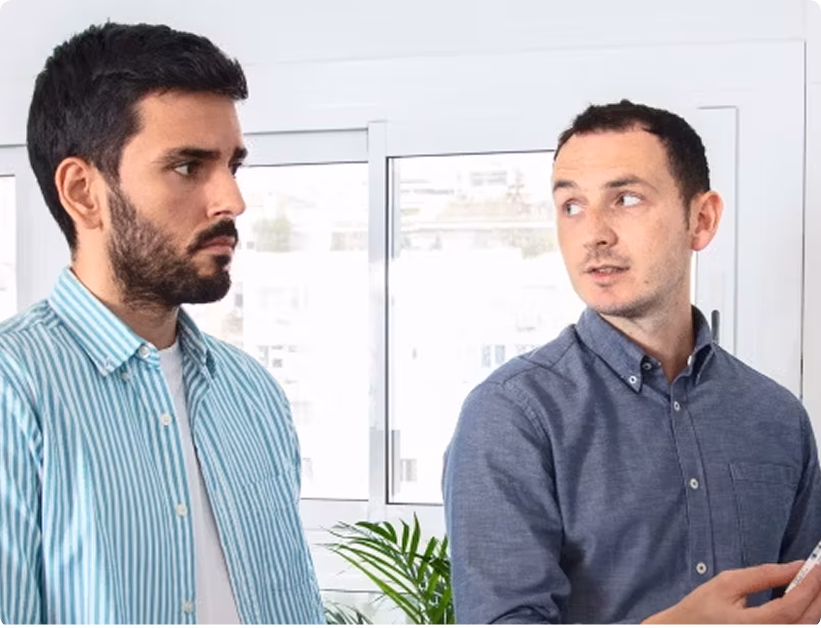 Man in blue shirt talking to another man in striped shirt, in a well-lit office setting