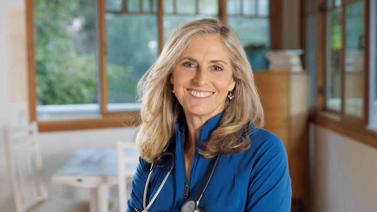 Delaney Ruston MD, smiling to camera in front of wide windows with green tree foliage