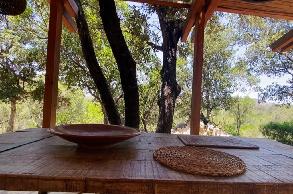 Bed with two striped pillows beside a window covered by sheer white curtains with sunlight streaming through.