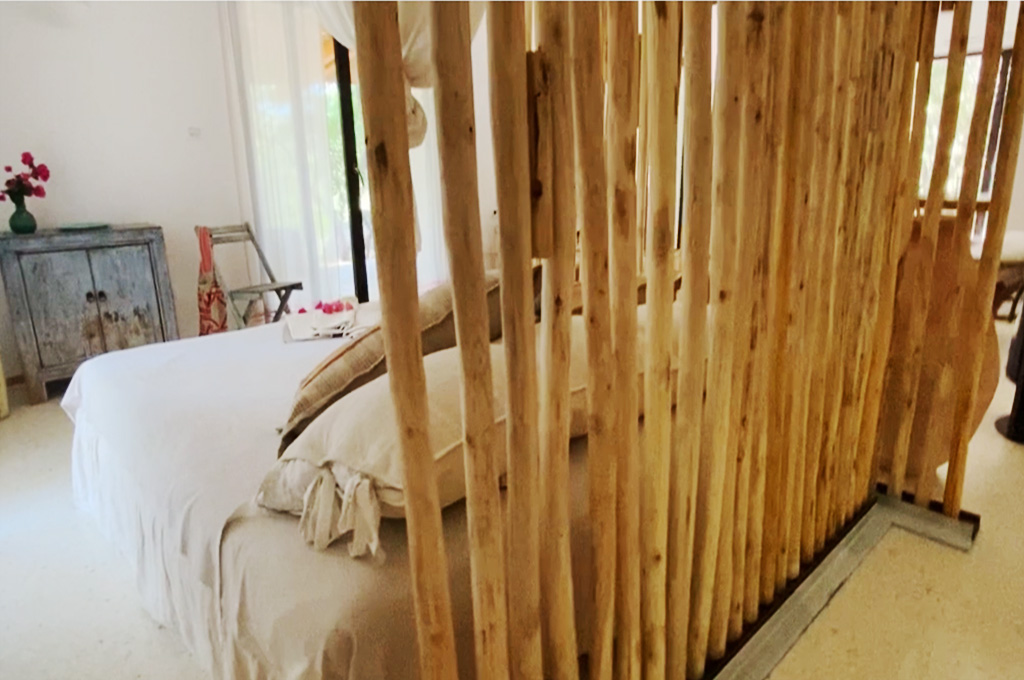 A woman in a pink and white dress slicing a tomato on a wooden cutting board outdoors next to a pot of lettuce and a stovetop with a pot of water.