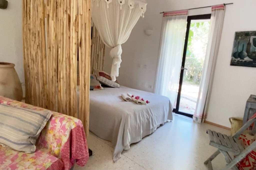 Cozy bedroom with a bed draped in white linens under a white canopy, a floral upholstered chair, and a glass door letting in natural light.