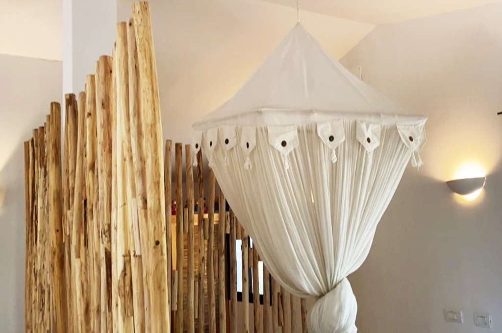 Woman in pink and white dress smiling while standing behind a wooden table set outdoors with plates, a bottle, and a cutting board with tomatoes.