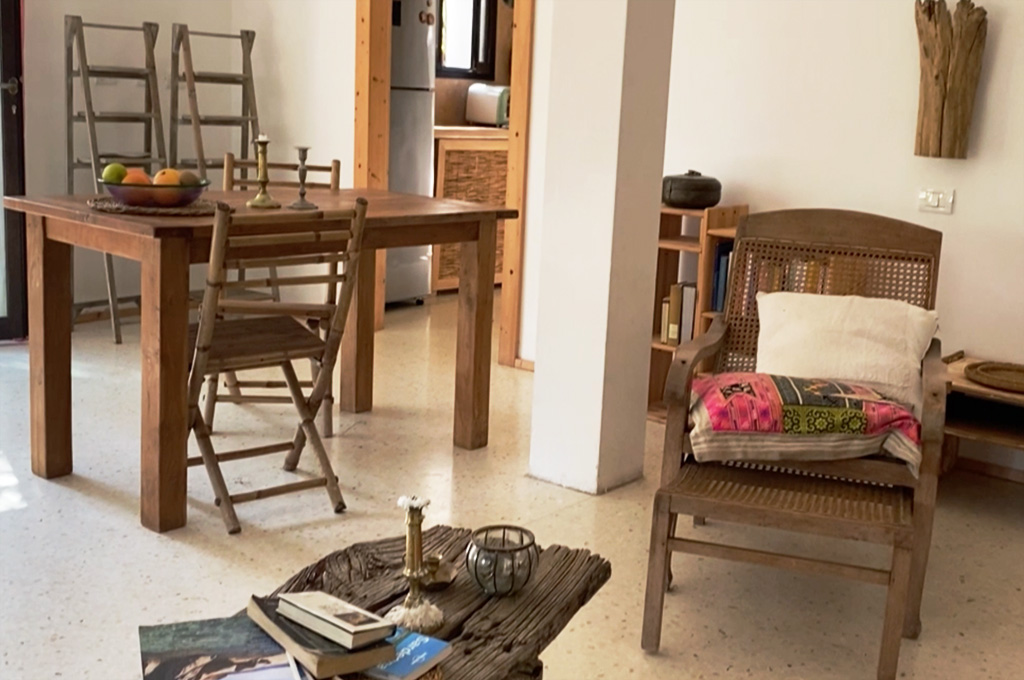 Cozy room with wooden table and chairs, a woven wooden armchair with colorful cushions, a rustic coffee table with books and candles, and a small bookshelf.