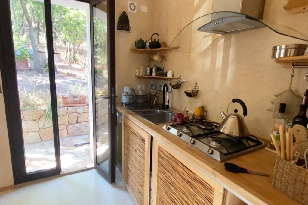 Cozy kitchen with a gas stove, kettle, sink, wooden cabinets, and an open glass door leading outside to a garden.