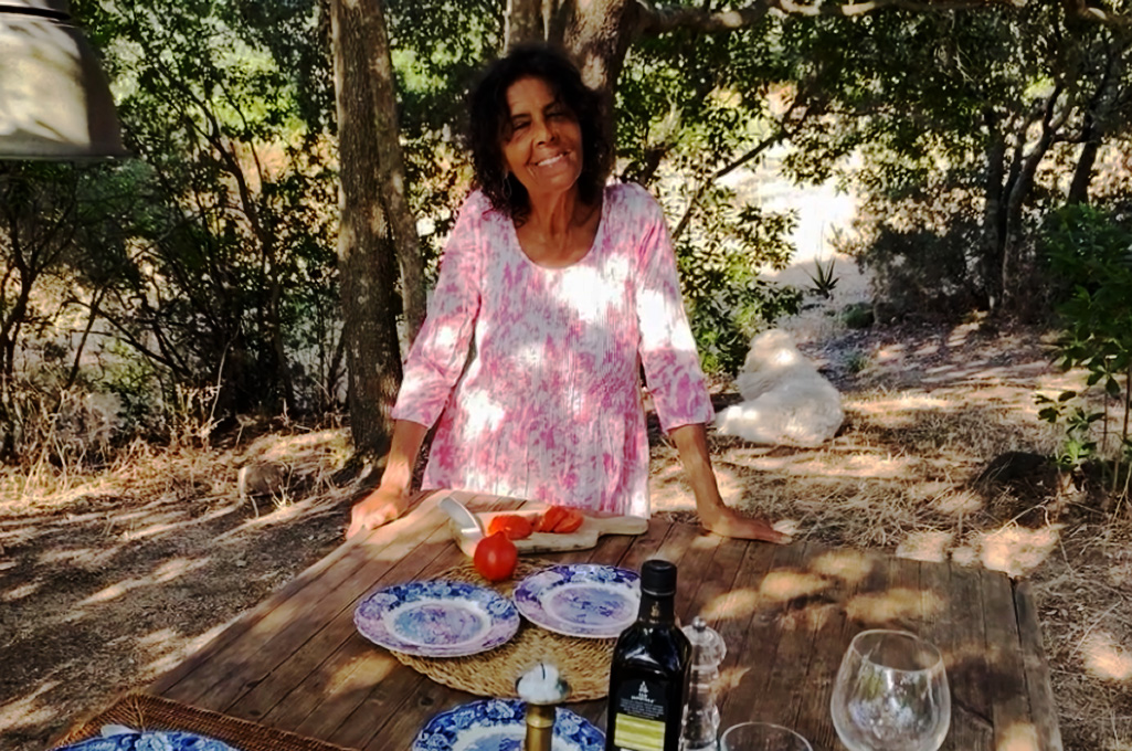 Woman in pink dress standing behind a wooden outdoor table set with plates, wine glasses, olive oil, and a cutting board with tomatoes.